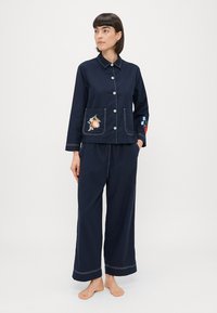 Tumši zilas kokvilnas pidžamu komplekts ar pogām augšdaļā, kas dekorēts ar ziedu izšuvumiem kabatās un piedurkņos; platām biksēm; kontrastējošām baltām šuvēm.
