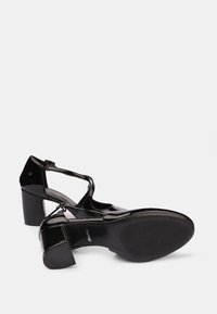 Chaussures en cuir verni noir à bout fermé, avec une bride ankle et un talon bloc. La semelle présente un motif nervuré pour une meilleure adhérence.