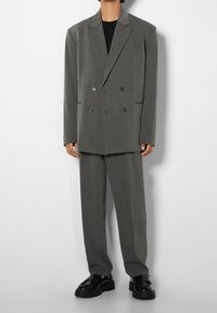 Person klädd i en överdimensionerad grå dubbelknäppt kostym med svarta loafers och en svart skjorta, stående framför en enkel vit bakgrund.