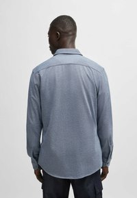 Camisa de manga larga en azul claro, de tejido texturizado. Cuenta con un cuello estándar y puños abotonados. Doquier curvo visible desde la vista posterior.