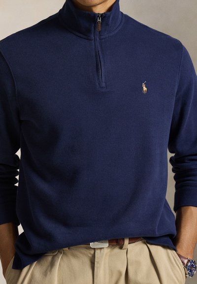 Tengerészkék negyed cipzáras pulóver kis hímzett pólo játékos logóval, bézs nadrággal és barna övvel viselve, kezek a zsebekben.