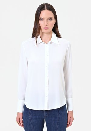Femme aux cheveux bruns et raides, portant une chemise blanche boutonnée et un jean bleu, debout devant un fond clair uni.