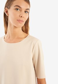 Top bej cu guler rotund, mâneci scurte și un material cu textură fină; prezintă un design subtil cu cusături minime.