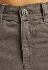 Jeans din denim maro cu o suprafață texturată, dotate cu un nasture decorativ din metal și bucle pentru curea, prezentate din talie în jos.