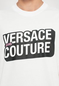 Balts kokvilnas T-krekls ar izteiksmīgu melnu tekstu, kas lasa "VERSACE JEANS COUTURE", ar rozā akcentu uz "JEANS". Vienkāršs dizains.