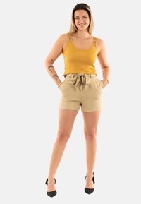 Débardeur jaune avec des bretelles décoratives, associé à un short beige taille haute avec une ceinture à nouer, porté avec des talons noirs.