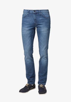 Jeans ajustados de hombre en azul con bolsillos frontales y trabillas para cinturón, combinados con zapatos casuales azul marino con cordones.