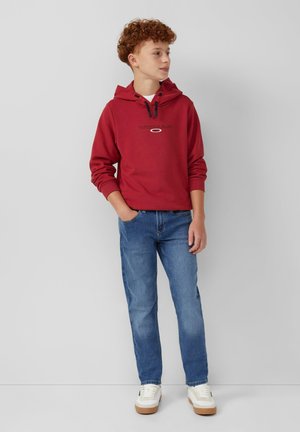 Rode hoodie met een trekkoord en logo tekst op de borst, gecombineerd met blauwe spijkerbroek en witte sneakers, op een lichtgrijze achtergrond.