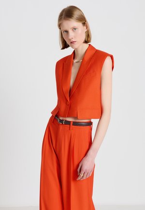 Femme aux cheveux blonds courts portant un blazer orange sans manches et un pantalon large taille haute assorti avec une ceinture noire, debout sur un fond blanc.