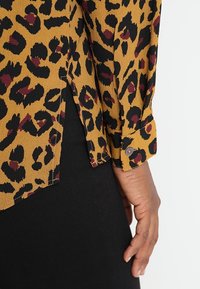 Blusa a fantasia leopardata gialla senape e nera, con polsini abbottonati e un'orlo leggermente curvo che rivela un indumento di base nero.