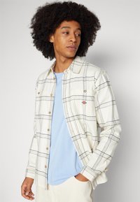 Weiß und blau kariertes Flanellhemd mit Knöpfen, das eine einzige Tasche und eine weiche Textur aufweist, getragen über einem hellblauen T-Shirt.