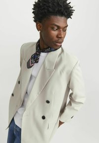 Blazer gris claro de doble botonadura con botones negros, que presenta solapas anchas y una textura suave. Combinado con una bufanda negra de estampado y una camisa blanca.