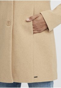 Beige Wolle Mantel mit einem Knopf, Seitentasche und einem kleinen schwarzen Logo-Tag nahe dem Saum. Hat eine weiche Textur.