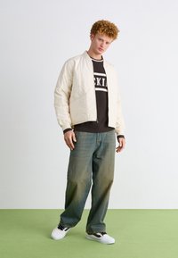 Hvid quiltet bomberjakke med ribbede manchetter over en sort sweater med hvid tekst. Kombineret med falmede blå vidde jeans og hvide sneakers.