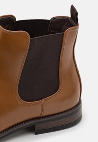 Botte Chelsea en cuir marron avec des panneaux latéraux élastiques, un bout arrondi et une semelle marron foncé avec un effet bois texturé.