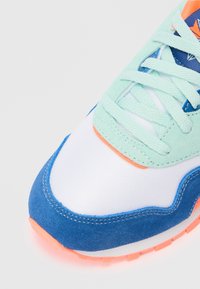 Acercamiento de la puntera de una zapatilla con gamuza azul, malla blanca, gamuza verde menta cerca de los cordones y suela de goma naranja con un patrón en el dibujo.