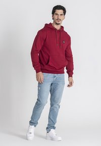 Červená bavlnená mikina s kapucňou, prednou kapsou, šnúrkami a malým logom. V kombinácii so zodratými svetlomodrými džínami a bielymi teniskami.