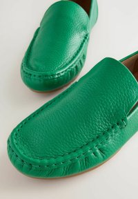 Chaussures slip-on en cuir vert avec un design cousu, un bout rond et une finition texturée. Les semelles sont plates et d'une couleur brun clair.