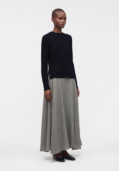 Femme portant un haut noir à manches longues et une jupe longue grise fluide, debout devant un fond blanc uni.