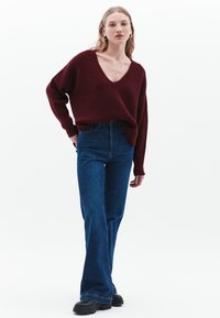 Bordeaux gebreide trui met een V-hals en een ruime pasvorm, gecombineerd met uitlopende blauwe denim jeans. De outfit bevat zwarte chunky laarzen.