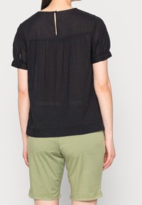 Blusa nera a maniche corte in tessuto testurizzato, con dettagli di pieghe e una piccola chiusura con bottone sul retro. Abbinata a pantaloni corto verdi.