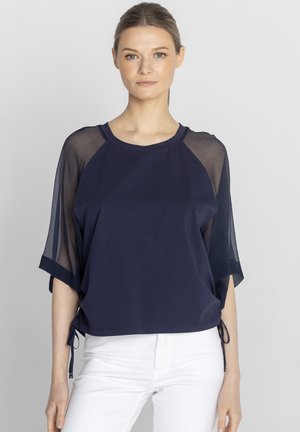 Kobieta w granatowej bluzce z przezroczystymi rękawami i wiązaniami, zestawiona z białymi spodniami, stoi na tle jednolitego jasnego tła.