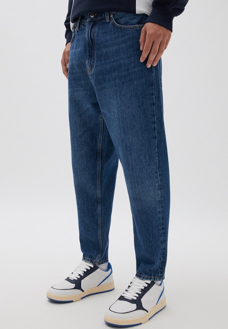 Personne portant un jean bleu foncé ample et des baskets blanches avec des détails noirs et bleus, debout sur un fond neutre.
