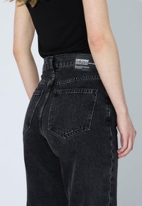 Persona che indossa jeans in denim neri a vita alta con tasche posteriori e un'etichetta del marchio DRDENIM visibile sulla vita.