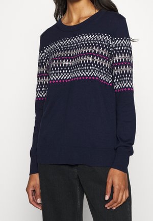 Mujer vistiendo un suéter azul marino con patrones geométricos blancos y rosas en el pecho y las mangas, combinado con pantalones negros.