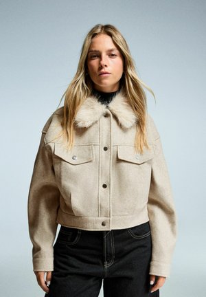 Korte beige jas gemaakt van een wolmix, met een nepbont kraag, knoopsluiting en twee borstzakken. Zwarte denim jeans zichtbaar daaronder.