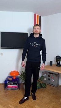 Μπλε σκούφο φούτερ με λευκό λογότυπο "Hilfiger", συνδυασμένο με μαύρο παντελόνι. Το μοντέλο στέκεται σε ένα δωμάτιο με πολύχρωμη διακόσμηση και έπιπλα.