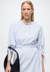 Φόρεμα με γαλάζιες και άσπρες ρίγες, με δέσιμο στον λαιμό, φουσκωτά μανίκια και ζώνη στη μέση, σε συνδυασμό με μαύρη τσάντα. Λείο ύφασμα.