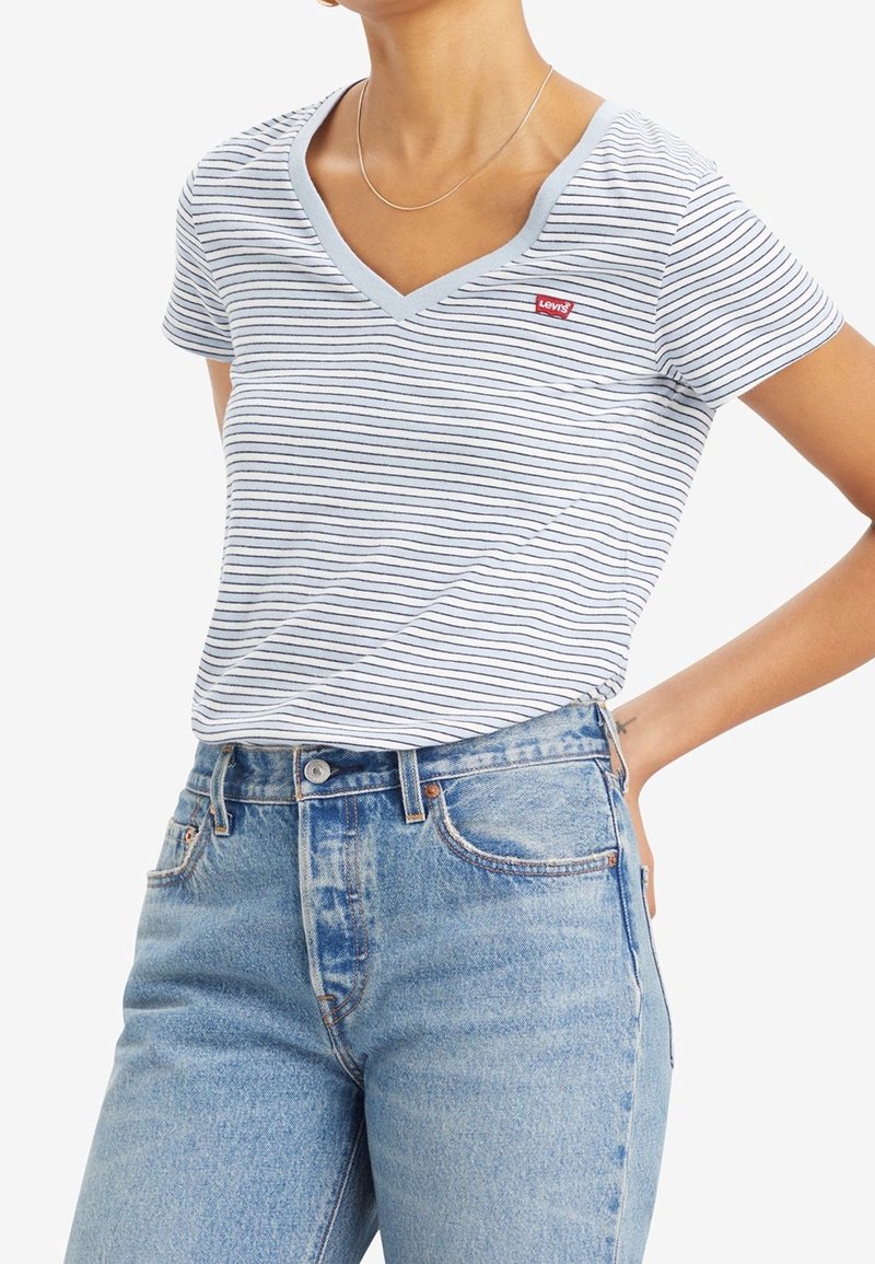 Personne portant un T-shirt Levi’s blanc et bleu marine à rayures avec col en V, rentré dans un jean taille haute bleu clair avec un collier en argent.