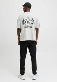 Grå t-shirt med korte ærmer og sort grafikdesign, der viser engle og tekst på ryggen, parret med sorte bukser og hvide sneakers.
