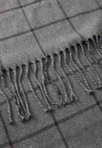Sciarpa grigia realizzata in materiale morbido, caratterizzata da un motivo a quadri nero e bordi frangiati, che ne esaltano la texture e il design.
