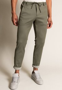 Olivově zelené tepláky vyrobené z jemné látky s elastickým pasem, nastavitelnou šňůrkou a zúženými lemy. Minimalistický design.