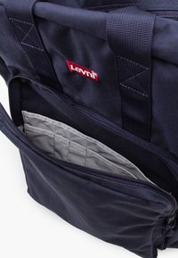 Tamsiai mėlynas drobinis kuprinė su paminkštintu pilku vidumi, pristatantis priekinę užtrauktuko kišenę ir tvirtas juodas diržus. Raudonas Levi's logotipas viršutinėje dalyje.