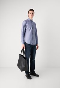 Camicia a maniche lunghe azzurra con motivo, jeans blu scuri, scarpe nere e una grande borsa a tracolla nera in pelle. Sfondo neutro.