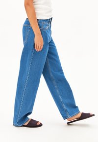 A person walks in blue wide-leg jeans and black slide sandals, showcasing a casual outfit against a plain white background.