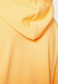 Sudadera amarilla con una textura suave, con una capucha suave y redondeada y un ajuste ceñido. La tela parece ligera y casual.