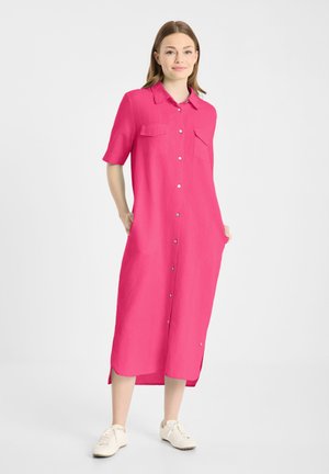 Kobieta stojąca w jaskrawo różowej sukience midi na guziki z krótkimi rękawami i białych tenisówkach, ręce w kieszeniach, na jednolitym białym tle.