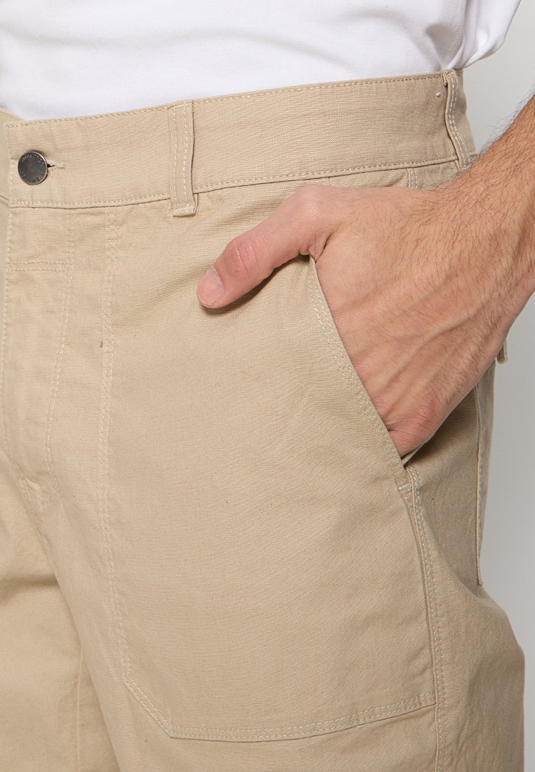 Shorts cargo en coton beige avec une main dans la poche latérale, dotés d'un bouton en métal et de coutures ajustées pour un look fitted.