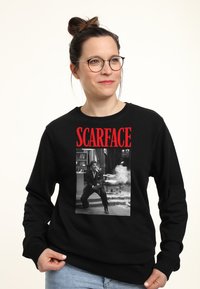 Felpa nera con una grafica di una scena del film in scala di grigi, con "SCARFACE" in caratteri rossi in grassetto sopra l'immagine.