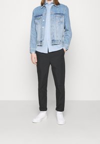 Giacca di jeans azzurri sopra una camicia azzurra con bottoni, abbinata a pantaloni neri e sneakers bianche. La giacca presenta due tasche sul petto.