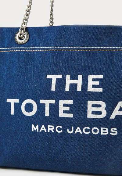 Denim auduma soma ar baltu tekstu "TOTE BAG" un "MARC JACOBS." Ietver ķēdes rokas un kontrastējošu šuvumu.