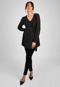 Svart glittrig graviditetströja med V-ringning, långa ärmar och figursydd midja, kombinerad med svarta leggings och spetsiga svarta klackar.