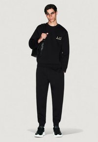 Joven vestido con sudadera negra y pantalones de chandal, llevando una mochila negra sobre un hombro, de pie contra un fondo claro y sencillo.