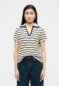 Γυναίκα με πλεκτό τοπ κοντομάνικο σε κρεμ και μαύρες ρίγες με V-λαιμόκοψη και σκούρο μπλε τζιν, στέκεται μπροστά σε απλό φόντο.