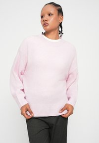 Ροζ πλεκτό πουλόβερ με στρογγυλή λαιμόκοψη, χαμηλούς ώμους και ριμπ τελείωμα. Φορεμένο πάνω από άσπρη μπλούζα, συνδυασμένο με σκούρα γκρι παντελόνι.