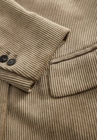 Beige korduroy sako s rukávy a kapsou s dvěma tmavými knoflíky, ukazující žebrovanou texturu a detaily složené tkaniny.
